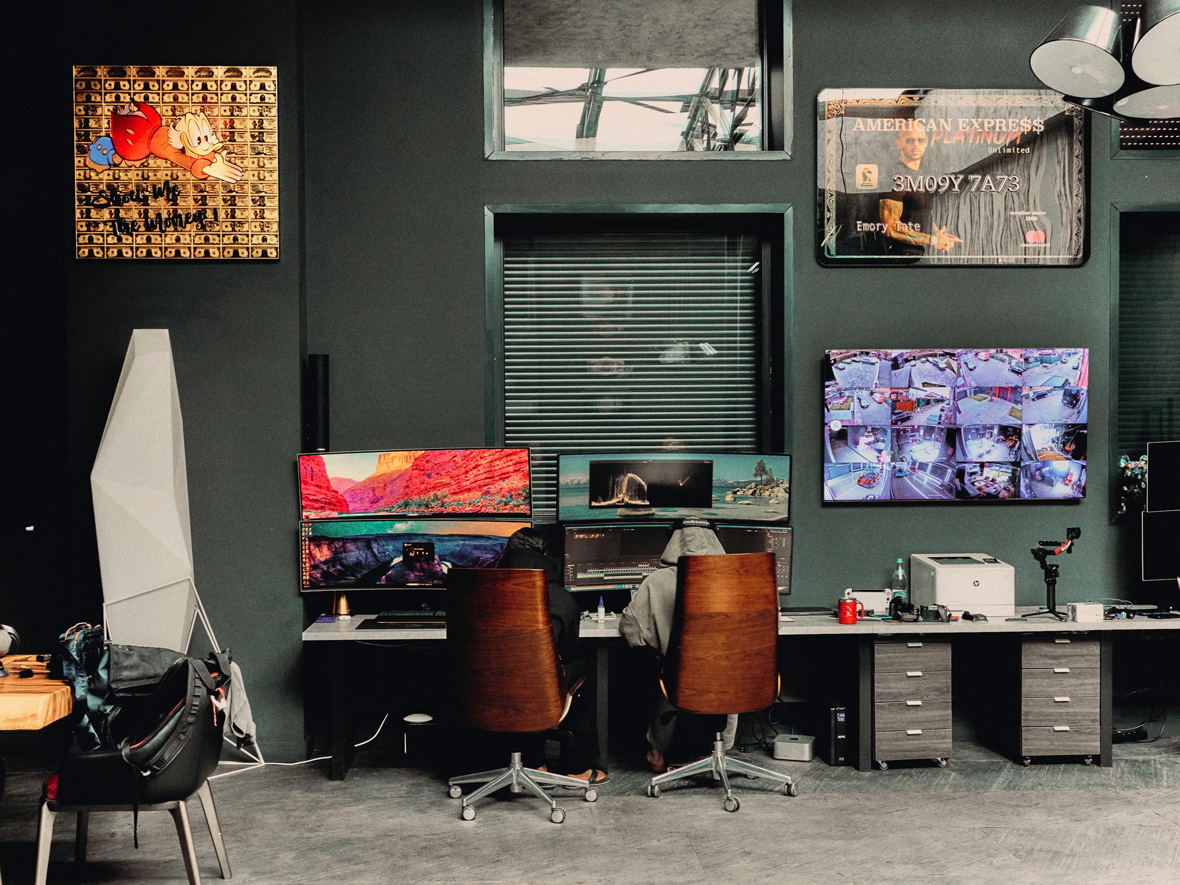 Two people in hoodies sit at a long desk of monitors in a green-walled studio, with framed money-themed art and a bank of security-camera feeds on the wall.