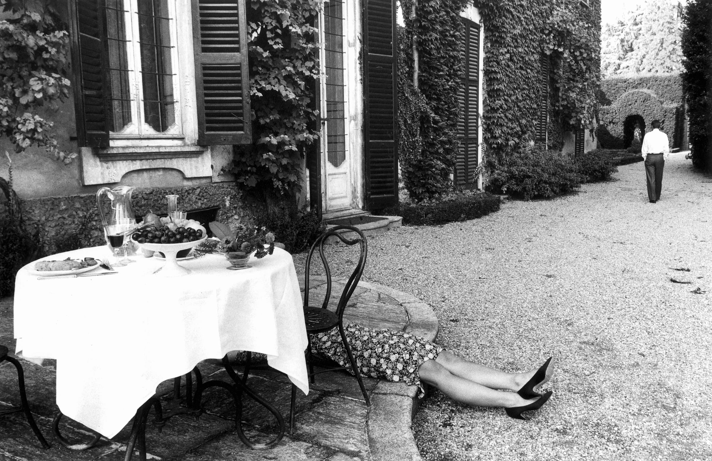 A black-and-white photograph of a patio table set with grapes and wine, a woman lying face-down behind a chair, and a man walking away down a gravel path.