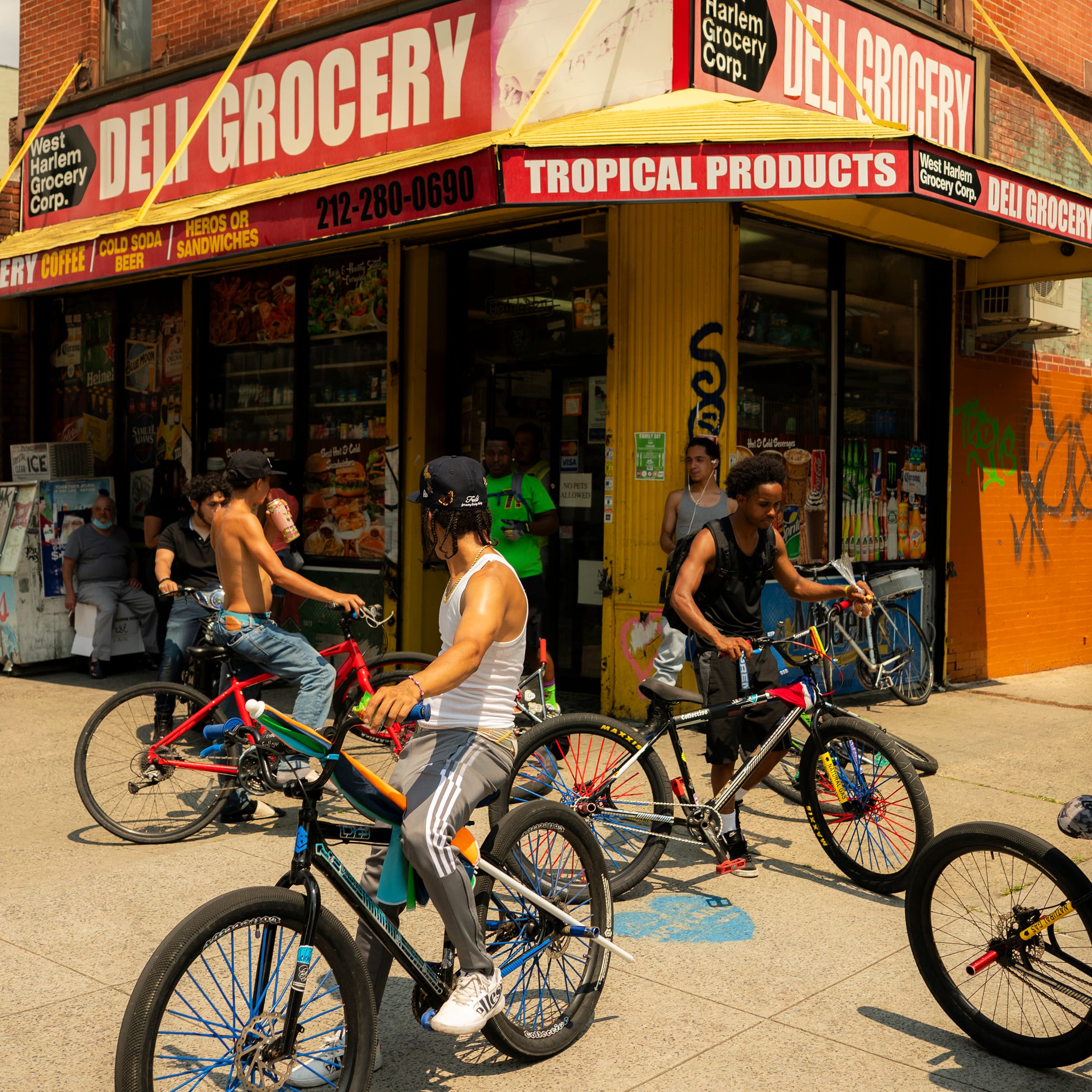 Young men with bicycles gather on a sunny Harlem sidewalk in front of a yellow deli grocery with red signage.
