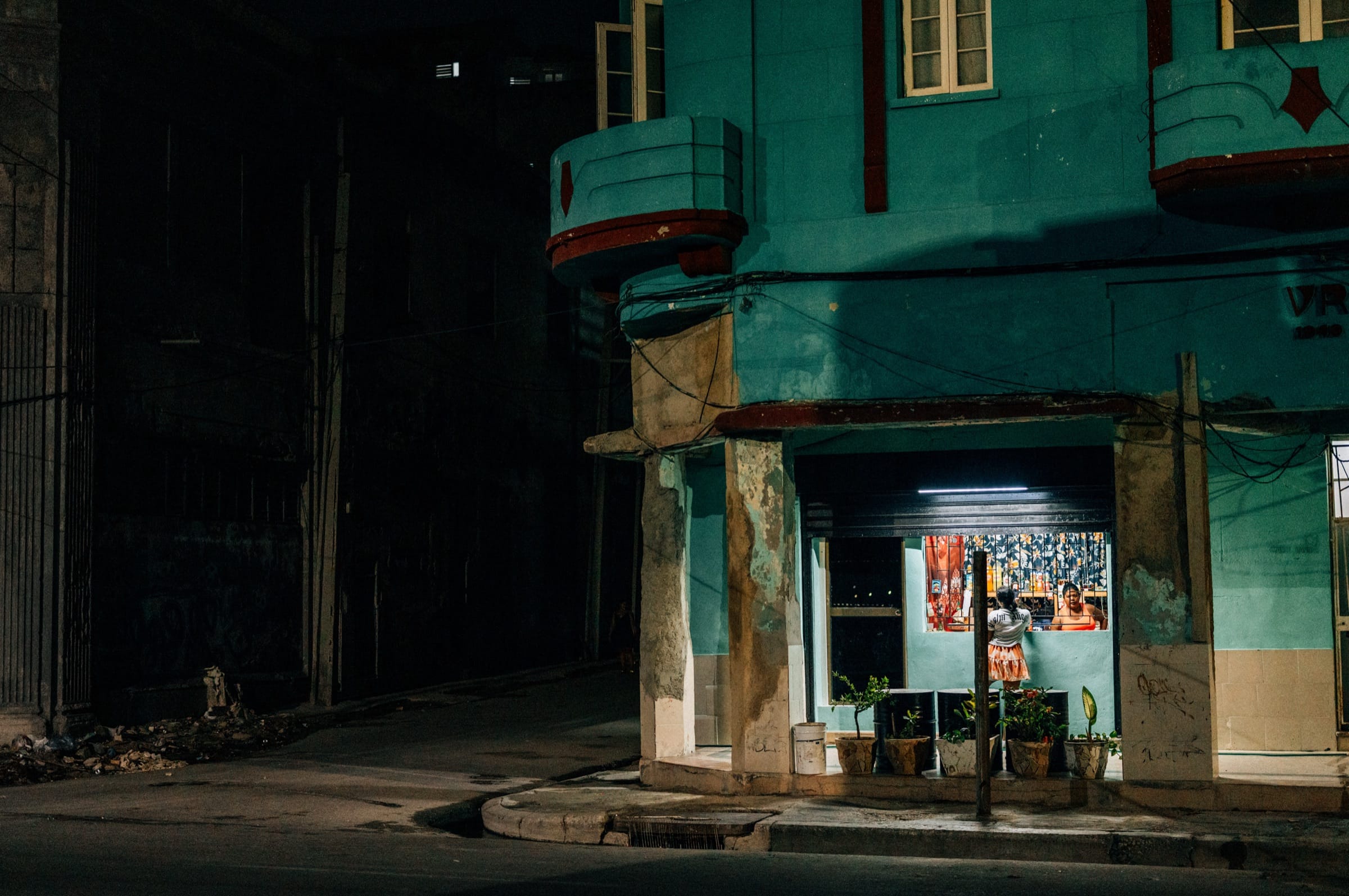 On a dark Havana street corner, a lit doorway in a turquoise building frames a small figure standing at a counter.