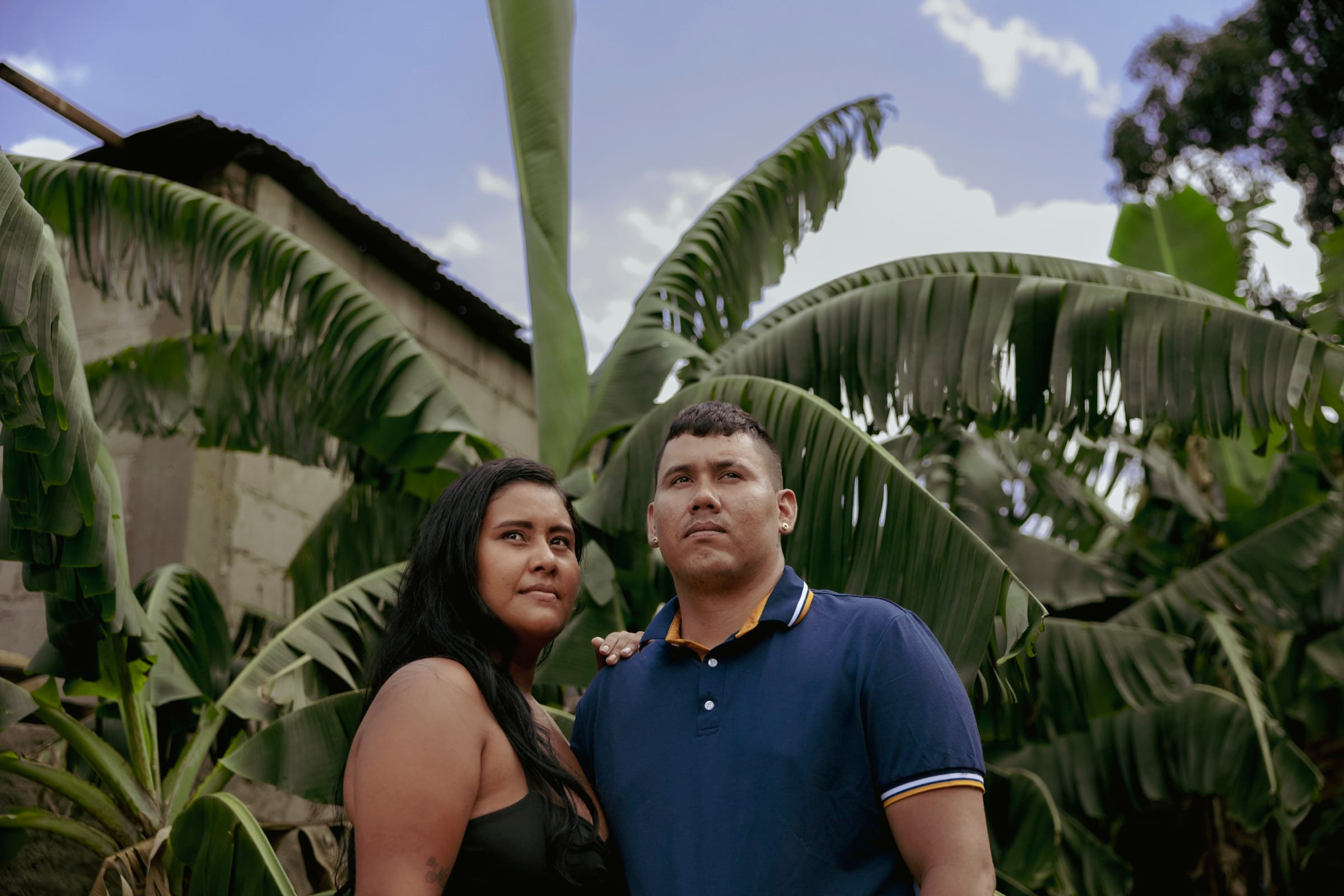 A woman and a man stand close together in front of broad banana leaves, both looking past the camera under a bright sky.