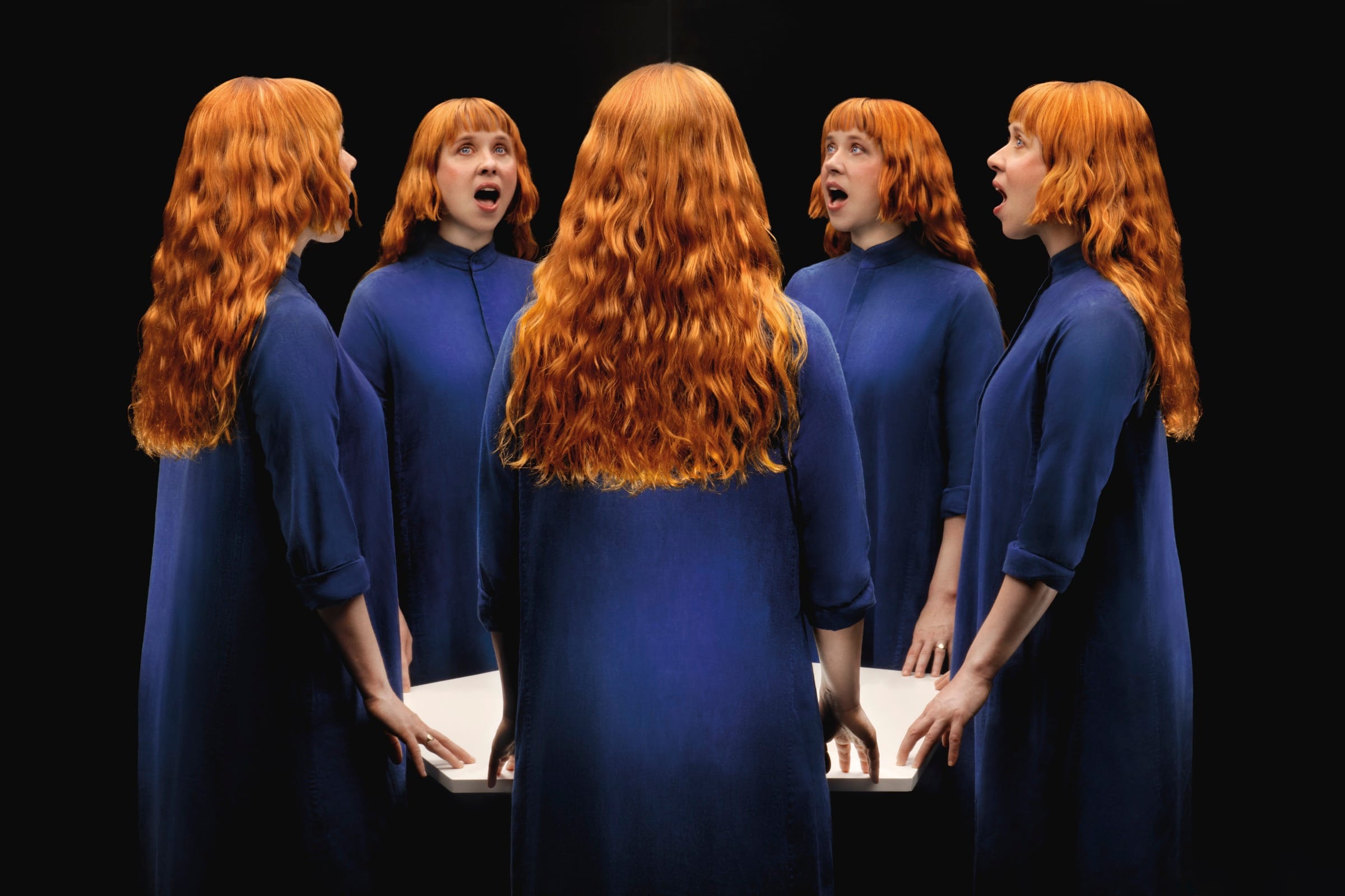 Five copies of the same red-haired woman in a blue robe stand around a white table against black, each singing or turned a different way.