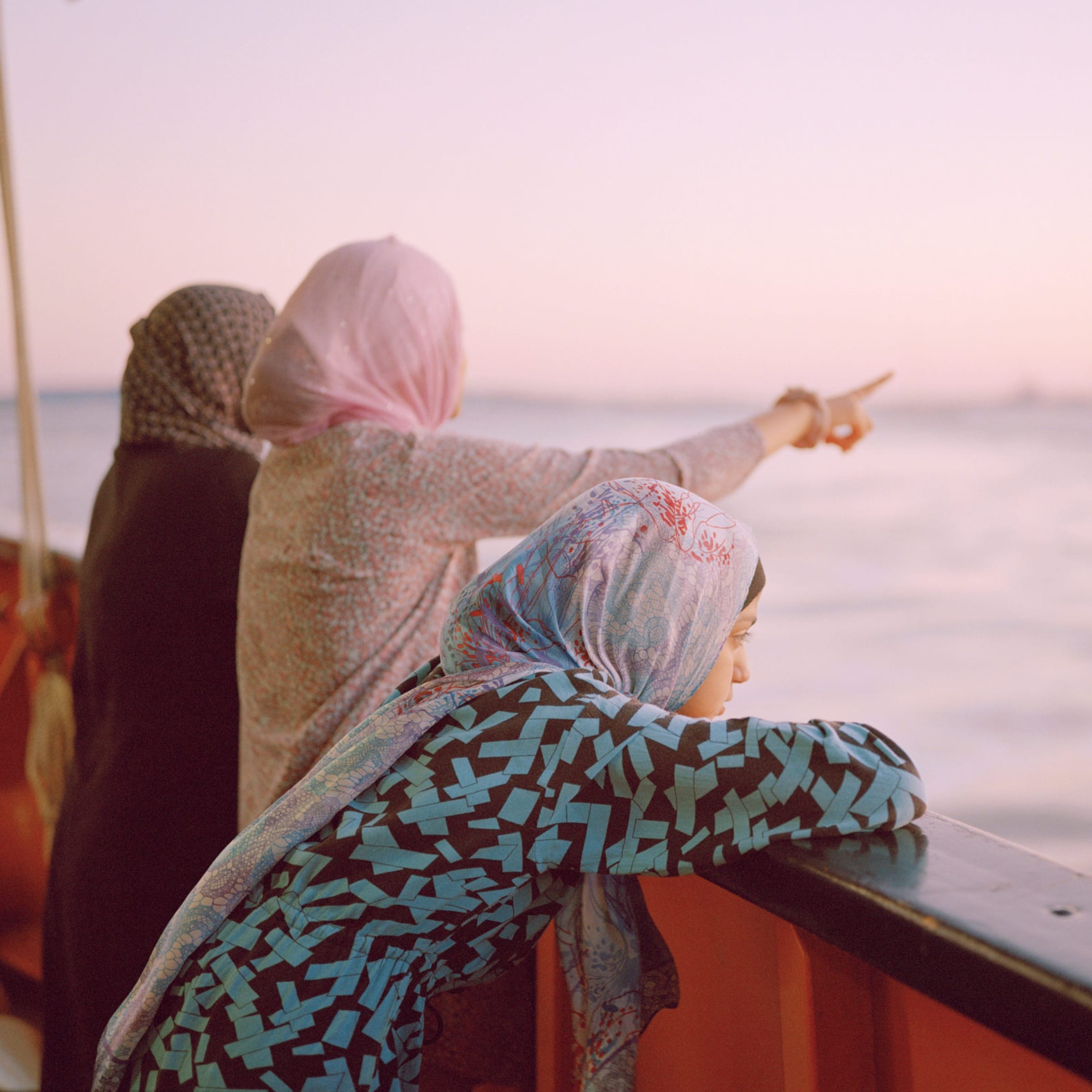 Three women in patterned head scarves lean on a boat rail at sunset, one pointing out across pale water.