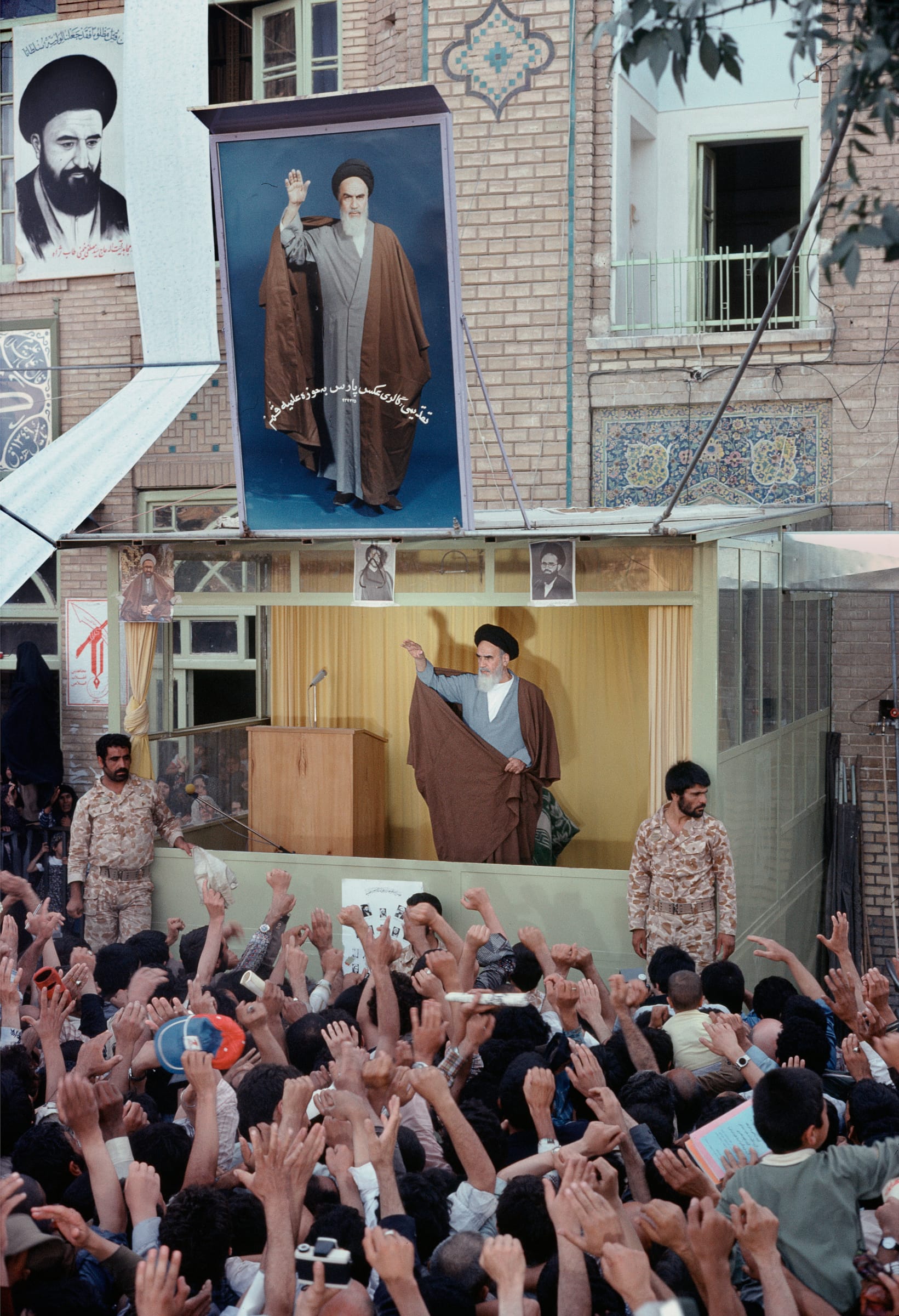 A crowd raises their arms toward a cleric speaking on an outdoor platform in Tehran, beneath a large painted portrait of Ayatollah Khomeini.