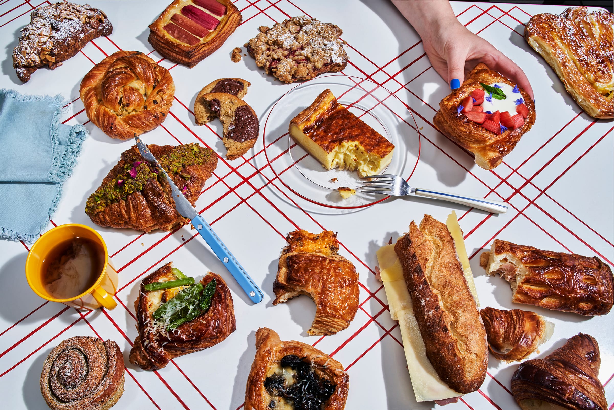 Pastries, a sandwich, cookies, and a coffee mug are scattered across a white table with a red grid, as a hand with blue nail polish reaches for a strawberry tart.