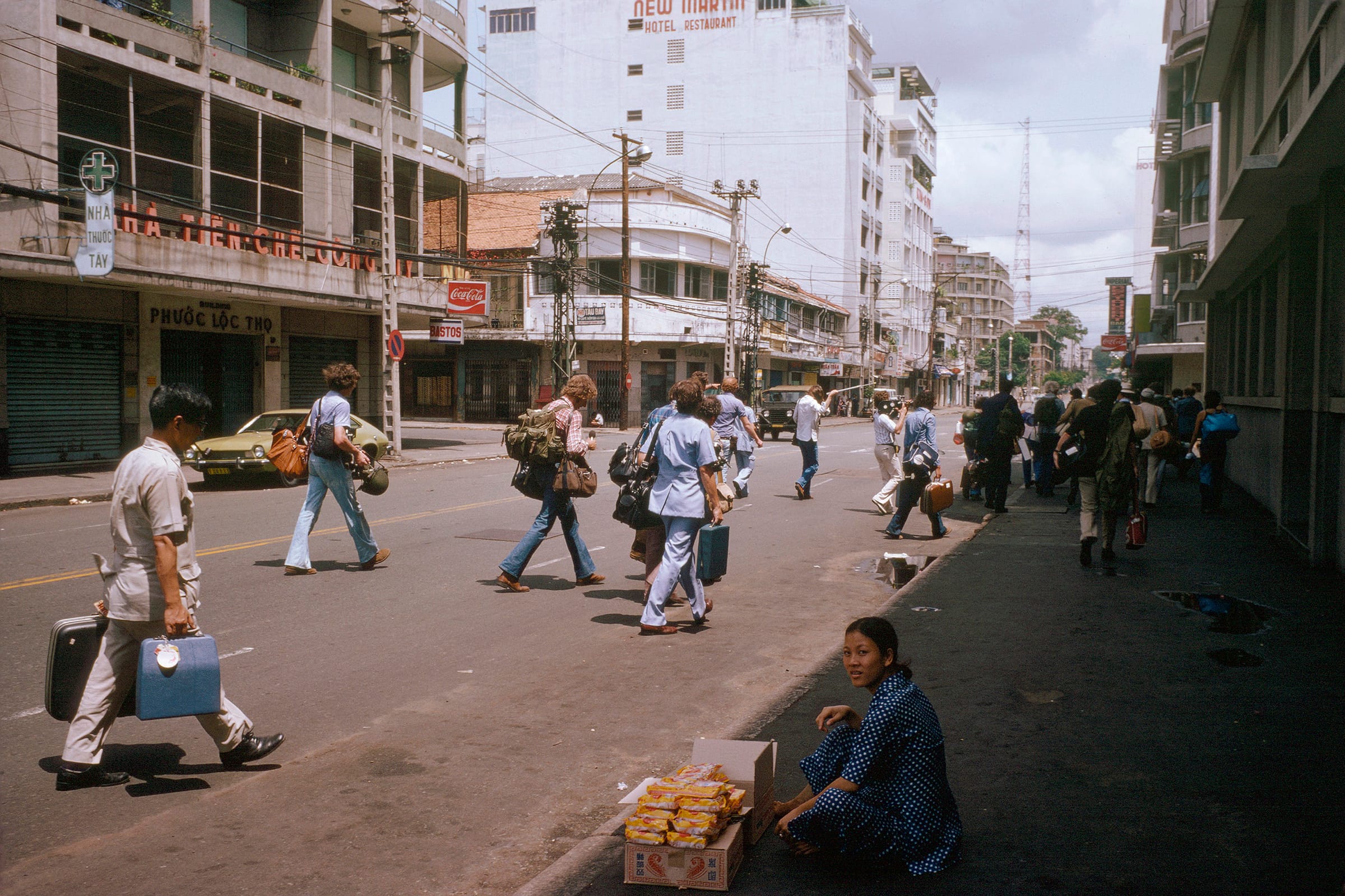 Westerners with luggage and camera gear cross a Saigon street lined with shop signs, while a woman in a polka-dot dress sits beside stacked boxes on the curb.