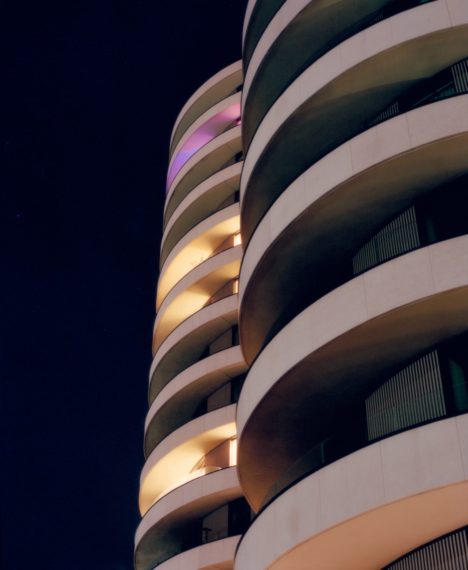 A curved apartment tower at night, its stacked balconies lit in bands of amber and violet against a black sky.