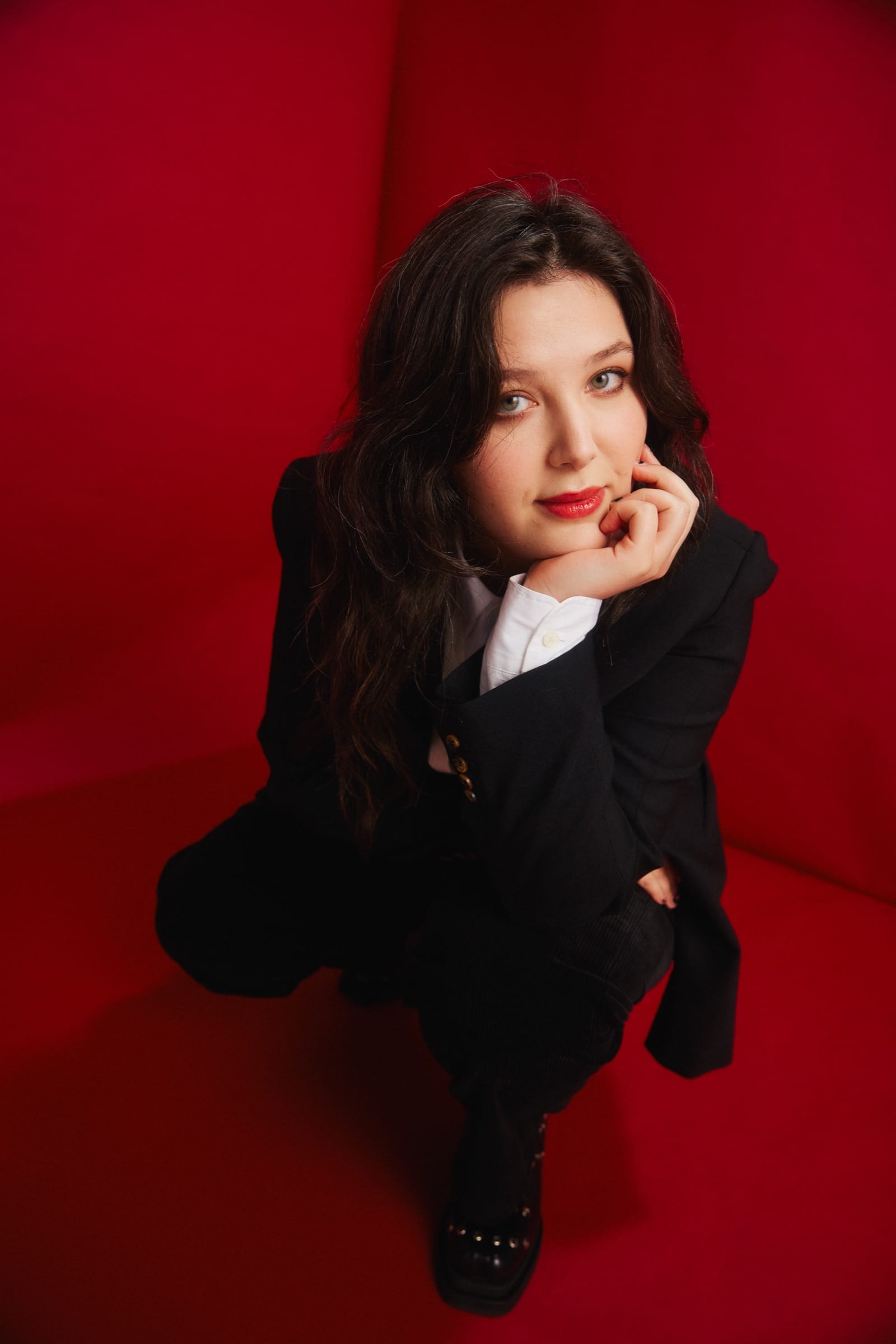 Lucy Dacus crouches in a black suit against a saturated red backdrop, chin resting on her hand, looking up past the camera.