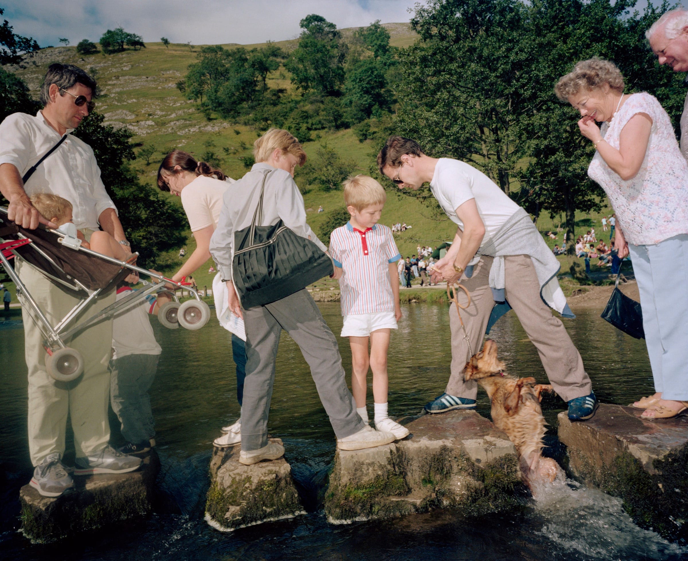 Holidaymakers pick their way across stepping stones in a shallow river, one man hauling a reluctant dog out of the water.