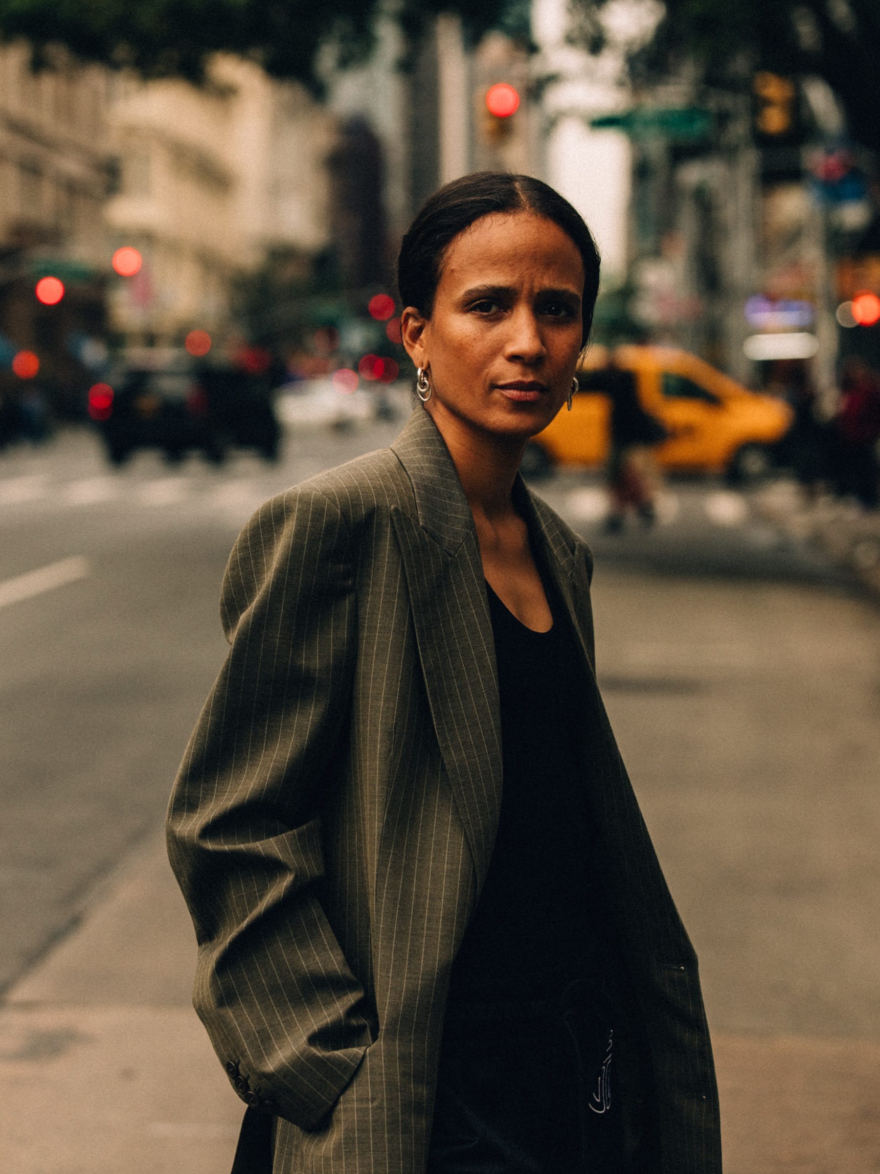The filmmaker Mati Diop stands in an oversized pinstriped blazer in the middle of a city street, traffic blurred behind her.