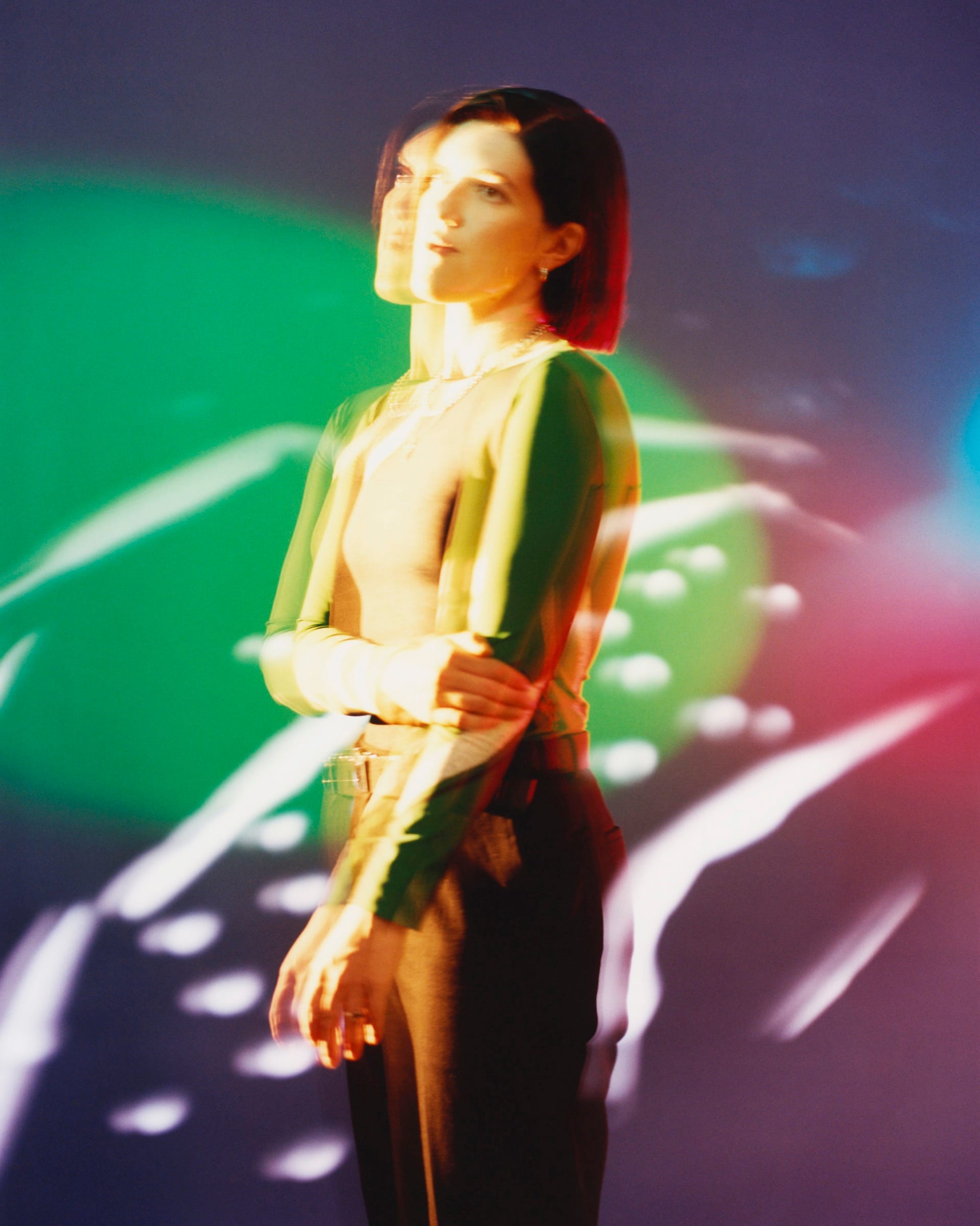 The musician Romy stands in a dark room, her figure doubled and streaked by green and pink projected light.