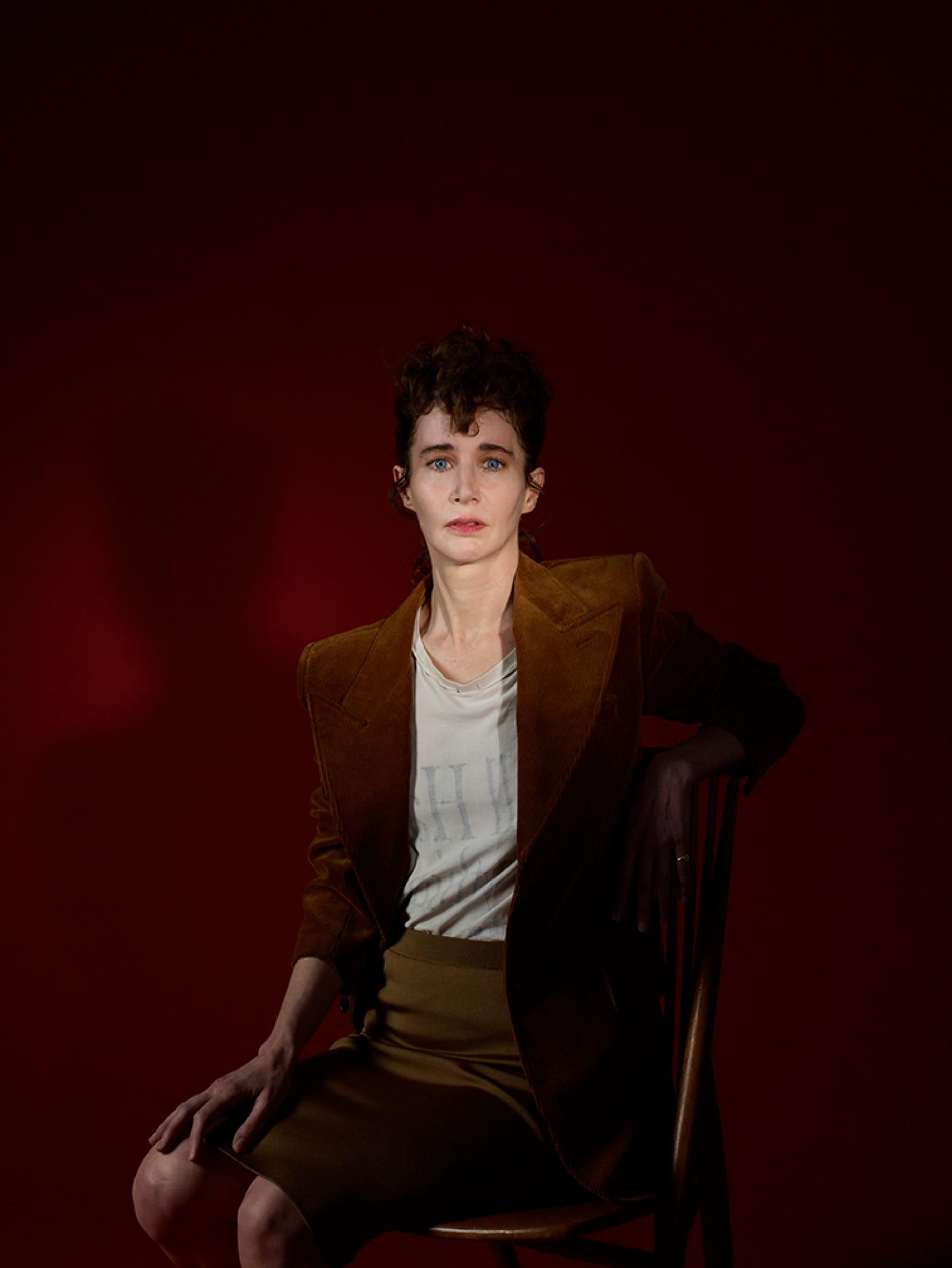 Miranda July sits sideways on a wooden chair in a brown suede blazer and skirt against a deep red backdrop.