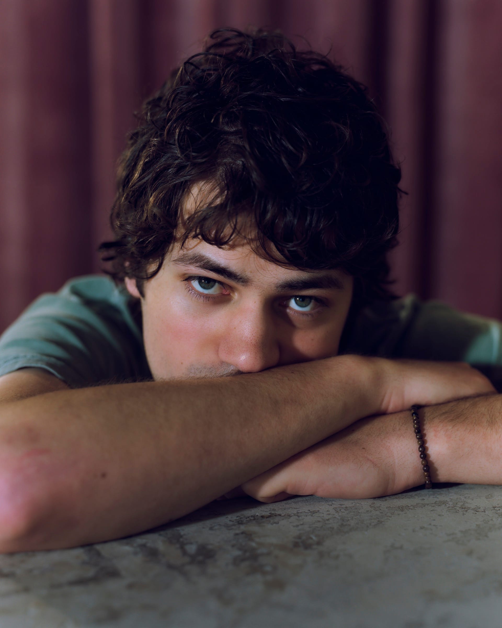 A young man with dark curly hair rests his chin on his folded arms atop a stone surface, looking straight at the camera.