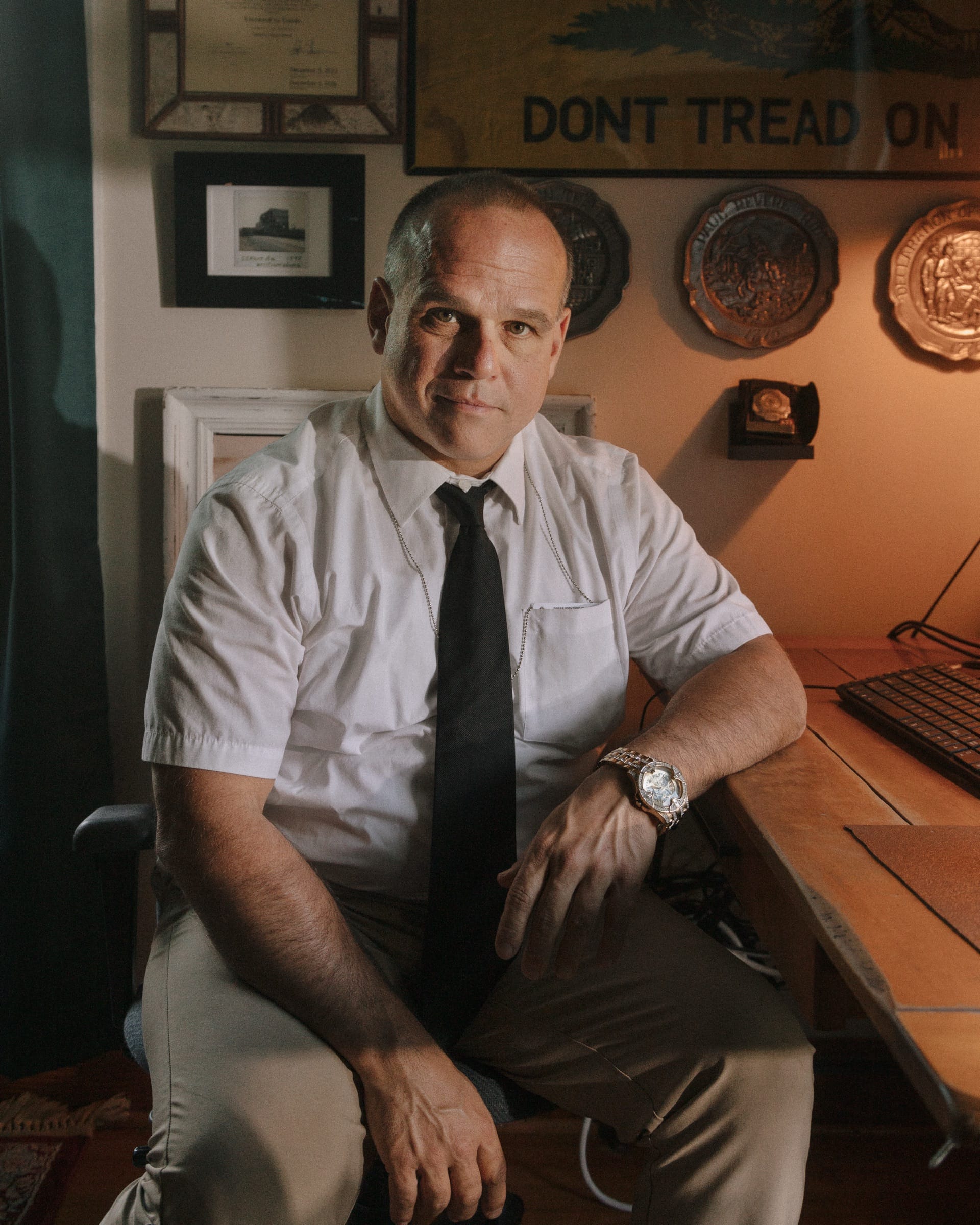 A man in a white shirt and black tie sits at a desk in a lamplit office hung with plaques, framed certificates and a Don't Tread On Me flag.