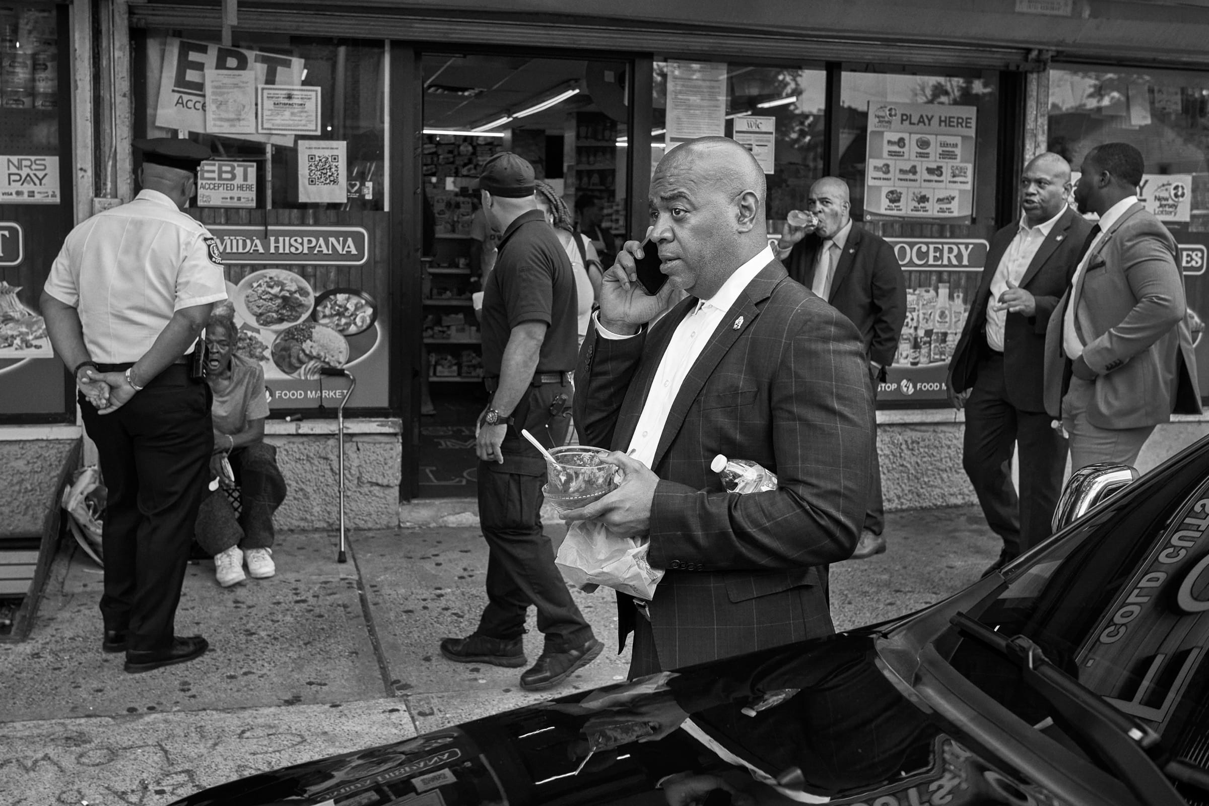 Black and white: Ras Baraka stands on a sidewalk outside a grocery, phone to his ear and a takeout container in his hand, aides nearby.