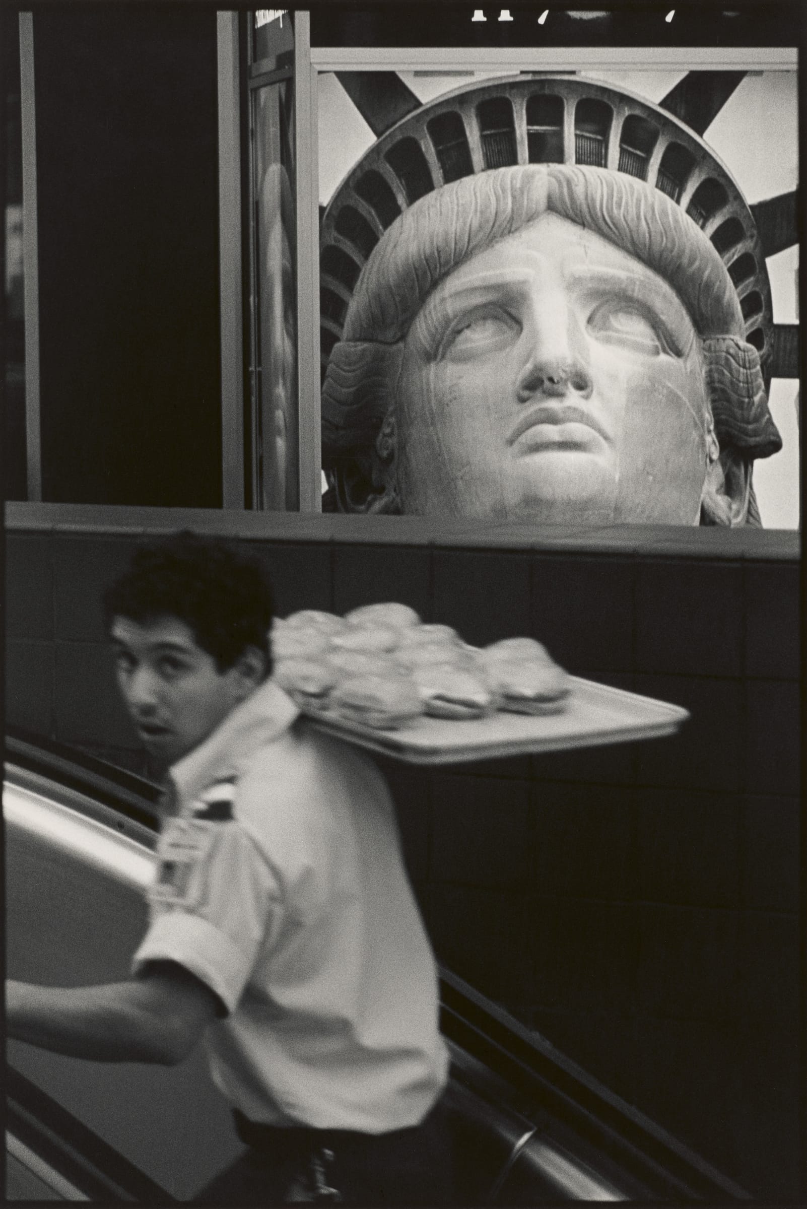 A black-and-white photograph of a man carrying a tray of sandwiches past a large image of the Statue of Liberty's face.