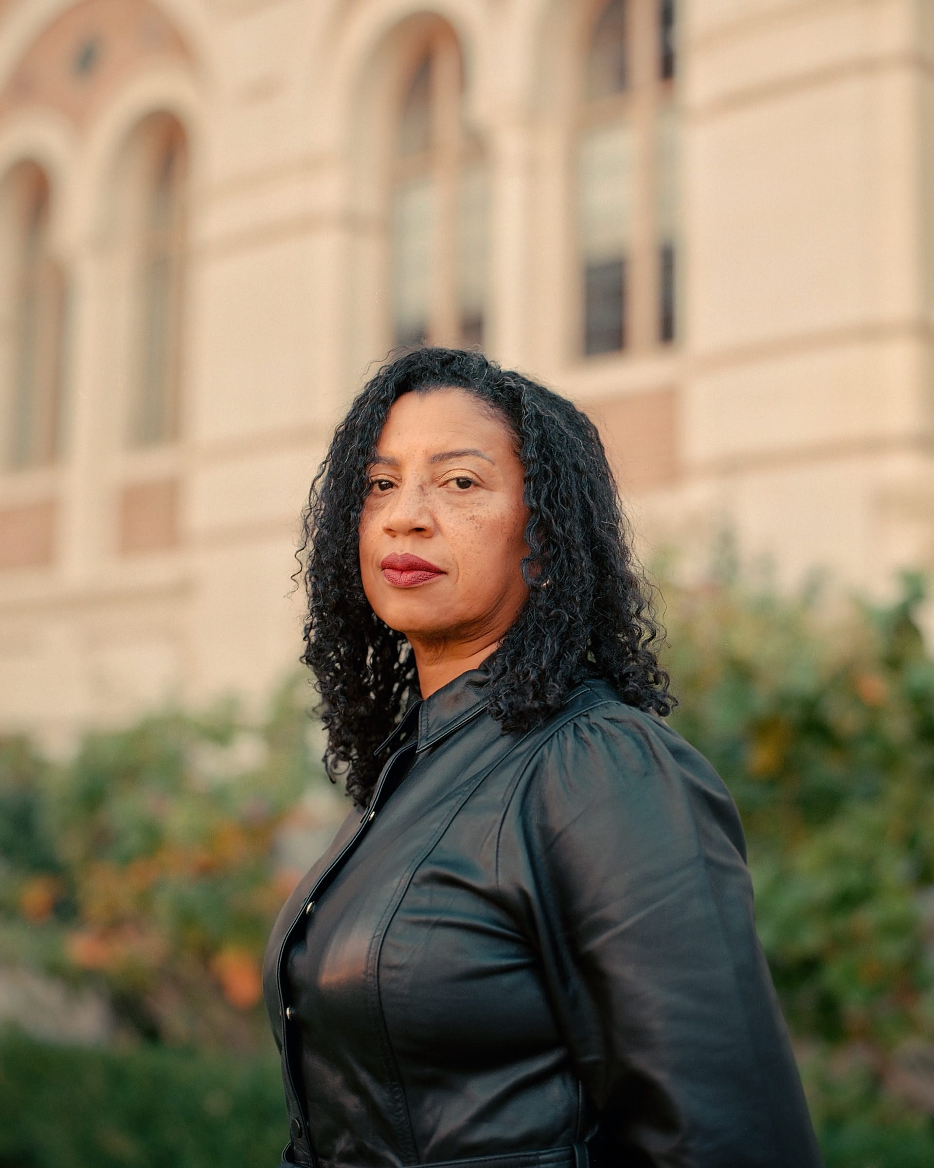 The poet Robin Coste Lewis, in a black leather jacket, turns to face the camera outside a pale stone building with arched windows.