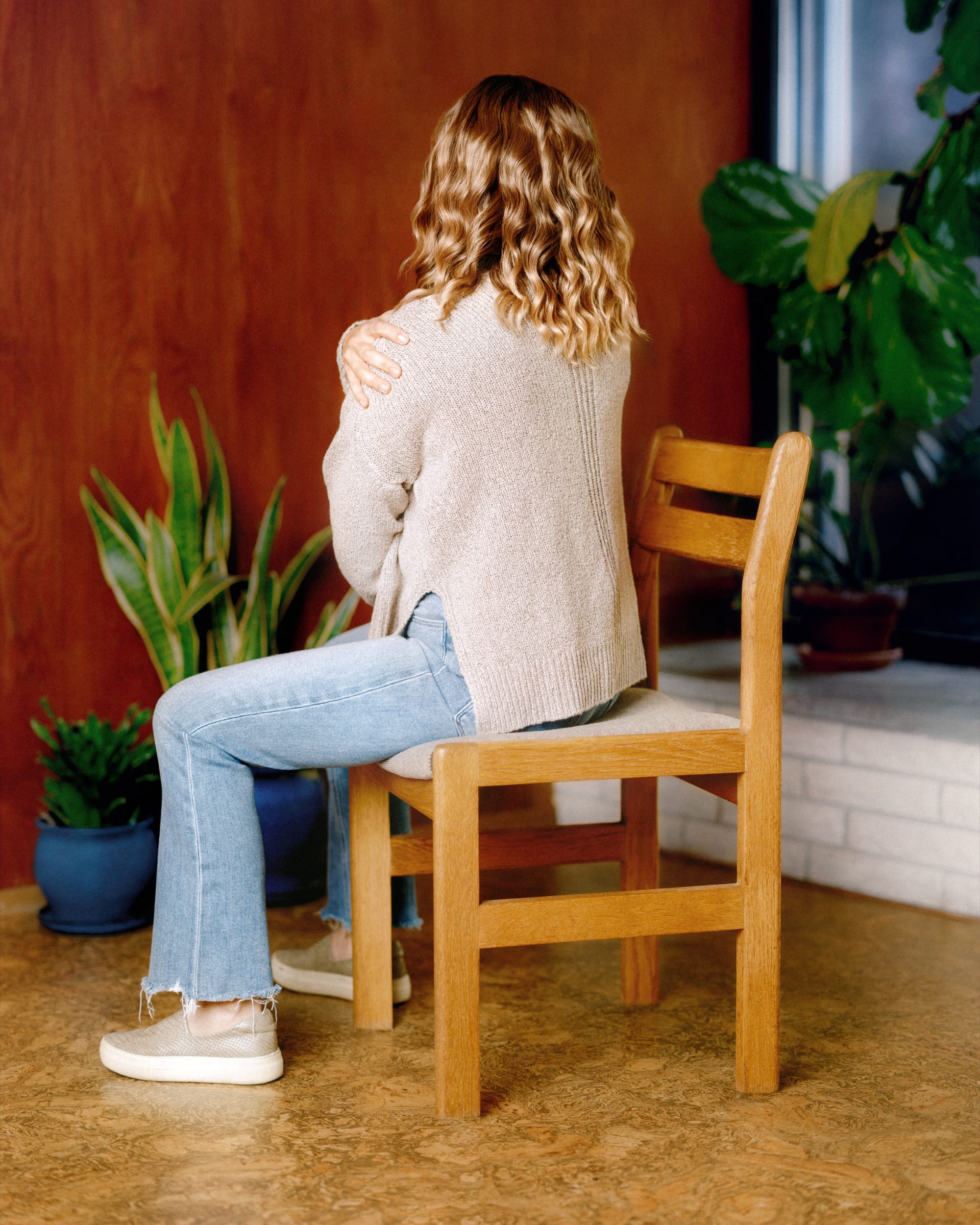 A woman sits on a wooden chair with her back to the camera, one hand gripping her shoulder, in a room with houseplants and a red wood wall.