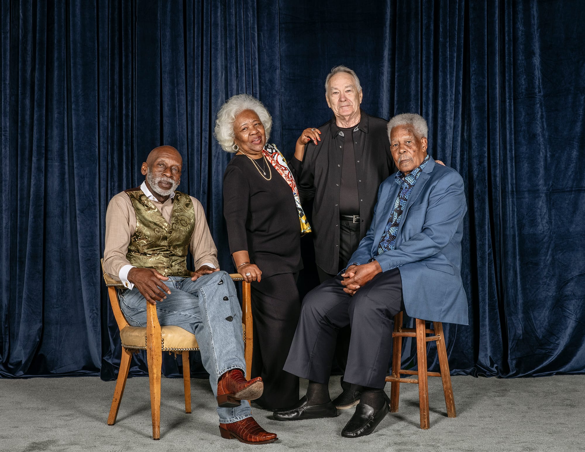 Four people pose for a studio portrait against a dark blue velvet curtain: two men seated in front, a woman and a man standing behind them.