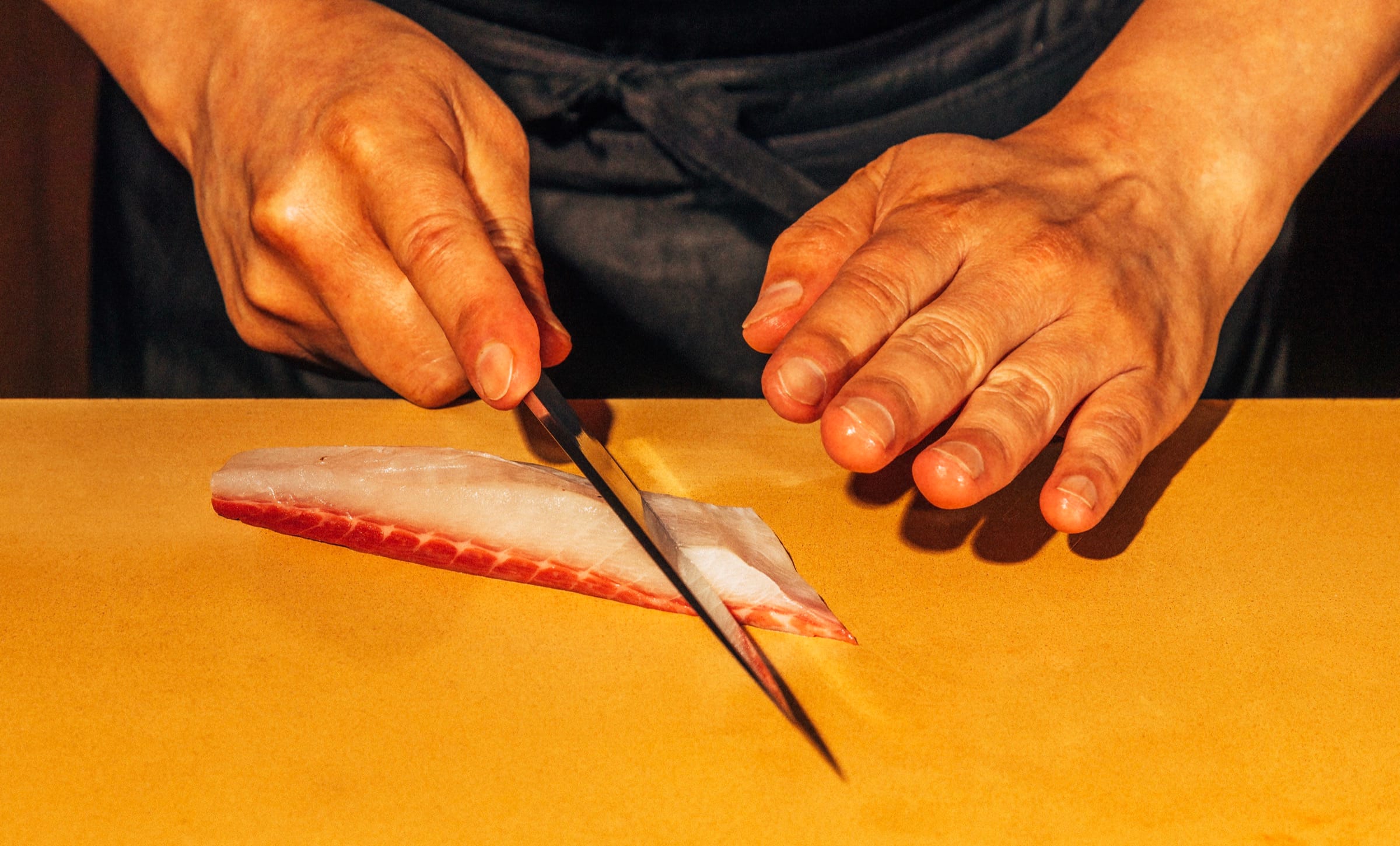 A chef's hands draw a long thin knife through a pale fillet of fish on a yellow cutting board.