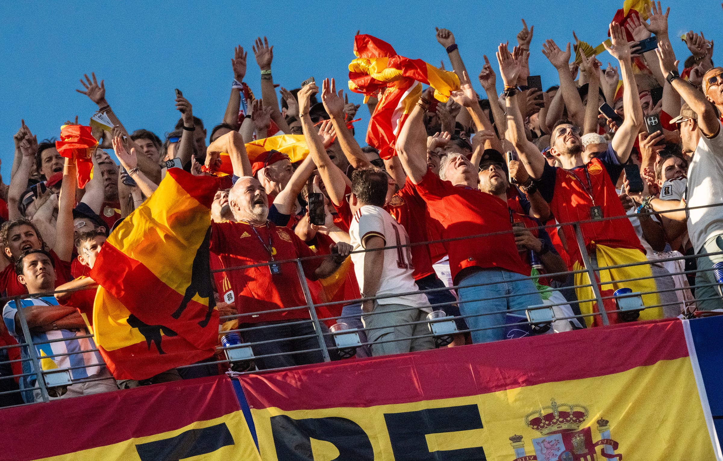 Spanish football fans in red and yellow cheer in a stadium stand, waving flags and phones under a clear blue sky.